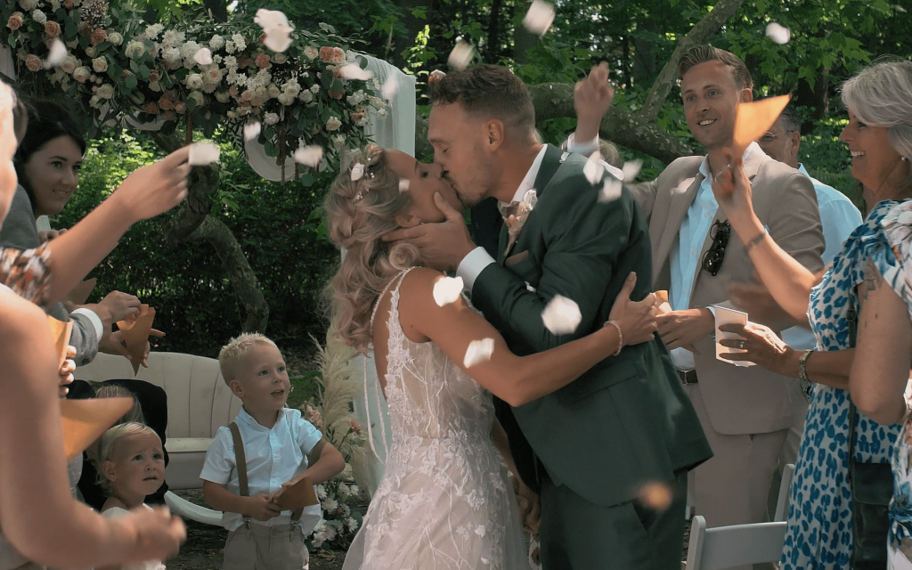 Bruidspaar kust tijdens ceremonie met bloemen en gasten.